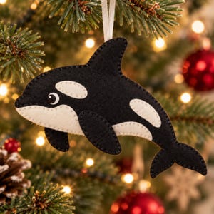 Op de afbeelding: Een zwart-witte orka-ornament van vilt met een gestikte rand en een wit lintje om op te hangen. De orka heeft een afgerond lichaam, een rugvin, borstvinnen en een staartvin, met een witte oogvlek en een wit gedeelte op de buik.