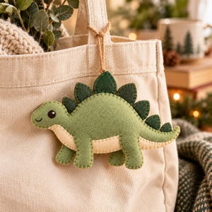 May include: A felt stegosaurus ornament in shades of light and dark green with a cream-colored underbelly, featuring visible stitching along the edges. The dinosaur is suspended by a thin brown twine string from a beige canvas tote bag.