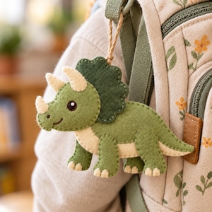 May include: A felt triceratops dinosaur keychain in shades of olive green and cream, featuring a dark green frill, three light-colored horns, and a brown twine loop for attachment, hanging from a beige backpack with a floral pattern.