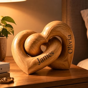 May include: A light-colored wooden sculpture consisting of two interlocking heart shapes. The name James is engraved on the left heart, and the name Olivia is engraved on the right heart. The sculpture features a visible wood grain pattern throughout.