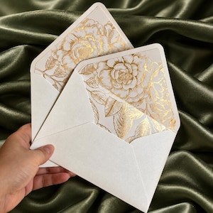 Puede incluir: Dos sobres de color crema con solapas puntiagudas, con un forro interior decorado con un patrón floral de color oro metálico de flores y hojas. El papel tiene un brillo sutil y los sobres están abiertos para mostrar el diseño interior.
