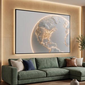 May include: A large rectangular wall art piece featuring a textured, relief-style map of North America. The landmass is rendered in shades of gray and white, with glowing golden lights highlighting the coastlines and urban areas.
