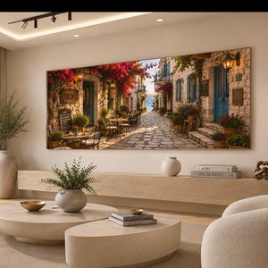 Op de afbeelding: Een grote rechthoekige wandkunstprint met een zonnige mediterrane kasseienstraat met stenen gebouwen, felroze bougainvillea-bloemen, blauwe deuren, terrasjes en een ver uitzicht op de zee onder een heldere lucht.