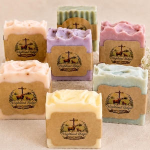May include: A collection of seven rectangular bars of soap in various pastel colors including pink, lavender, light blue, green, and cream. Each bar is wrapped in a brown paper label featuring a logo with a cow and the text Highland Ridge Soap Shop.