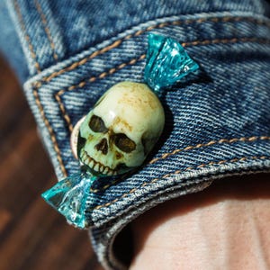 May include: A decorative pin shaped like a wrapped sweet with a translucent blue plastic wrapper and a realistic, off-white human skull in the centre, attached to the cuff of a blue denim jacket with visible orange stitching.