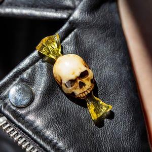May include: A pin shaped like a sweet wrapper with a central off-white human skull design. The ends of the wrapper are made of translucent yellow material, mimicking the appearance of twisted sweet packaging. The pin is attached to black leather.
