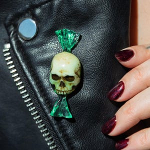 May include: A pin shaped like a sweet wrapper with a skull in the centre. The skull is off-white with dark shading in the eye sockets and nasal cavity. The wrapper ends are made of translucent, bright green material with a crinkled texture.
