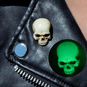 May include: A small, off-white skull pin with realistic brown shading in the eye sockets and nasal cavity. The skull is attached to a black leather jacket. An inset circle shows the same skull glowing with a bright, vibrant green light.