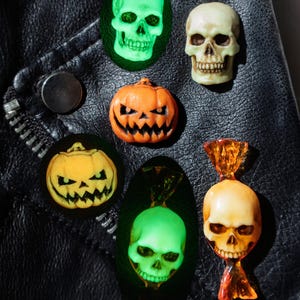 Könnte beinhalten: Eine Sammlung von Halloween-Anstecknadeln, bestehend aus einem realistischen beigen Totenkopf, einem orangefarbenen Kürbis, einem gelben Kürbis und einem durchscheinenden orangefarbenen bonbonförmigen Totenkopf sowie zwei leuchtend grünen Totenkopfmotiven.