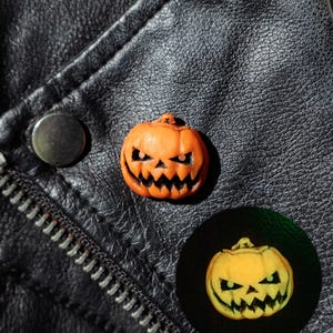 May include: A small orange jack-o'-lantern pin with a carved, jagged facial expression. The pin is shown in daylight and also in a circular inset that displays the pumpkin glowing with a bright yellow-green light in the dark.