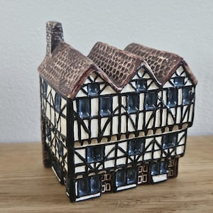 Pode incluir: Uma estatueta de casa em cerâmica com design estilo Tudor, apresentando estrutura de madeira preta, paredes cor de creme e um telhado castanho texturizado. A estrutura tem vários telhados, uma chaminé à esquerda e pequenas janelas.