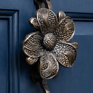 Könnte beinhalten: Ein bronzefarbener Türklopfer aus Metall in Form einer großen Blume mit sechs Blütenblättern. Die Blütenblätter haben eine texturierte, punktierte Oberfläche mit erhabenen, glatten, geschwungenen Linien. Die Mitte ist eine kreisförmige Scheibe.