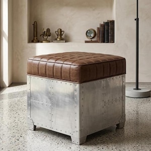May include: A square ottoman featuring a tufted brown leather top and a base constructed from riveted silver-toned metal panels. The base has a grid-like pattern of exposed rivets along the seams of the metallic plates, providing a structured look.
