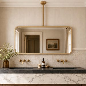 May include: A double vanity featuring a white marble front, a dark stone countertop, and a wooden base with a lower shelf. Above, a rectangular gold-framed mirror hangs from the ceiling. Gold faucets, a soap dispenser, and a woven basket are present.