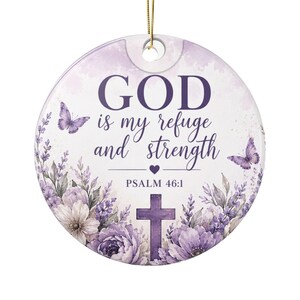 May include: A round ceramic ornament featuring a purple floral design with lavender flowers, two butterflies, and a cross. The text reads: GOD is my refuge and strength, PSALM 46:1. A small purple heart is centred below the text, and a gold string is attached.