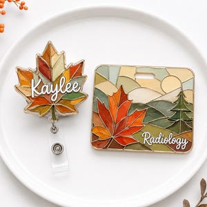 May include: A maple leaf-shaped badge reel with the name Kaylee in white script and a rectangular badge holder featuring a stained-glass style landscape with a large orange maple leaf, a green pine tree, and the word Radiology in white script.