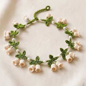 May include: A crochet necklace featuring a series of cream-coloured bell-shaped flowers and green leaves arranged along a thin green cord. The design is accented with small, round, white faux pearls placed between each floral cluster.