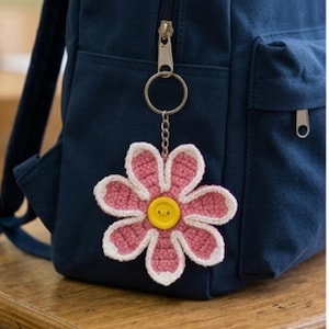 Pode incluir: Um porta-chaves de flor em croché com seis pétalas cor-de-rosa com bordas brancas e um botão amarelo brilhante no centro. A flor está presa a um anel e corrente de metal prateado, enganchada no fecho de uma mochila azul-marinho escura.