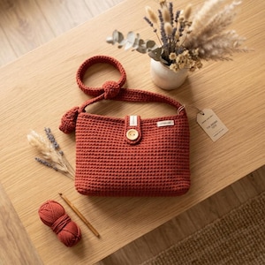 Könnte beinhalten: Eine rostfarbene Häkeltasche mit einem einzelnen Schulterriemen mit Knotendetail, einem zentralen Holzknopfverschluss und einem kleinen rechteckigen Etikett. Ein seitlich befestigtes Papieretikett trägt die Aufschrift The Sienna Tote. Ein Garnknäuel liegt daneben.