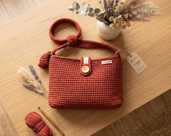 May include: A rust-colored crochet handbag featuring a single shoulder strap with a knot detail, a central wooden button closure, and a small rectangular label. A paper tag attached to the side reads The Sienna Tote. A ball of matching yarn is nearby.