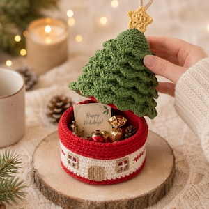 May include: A round, crocheted container with a red and white base featuring a house design, topped with a green, tiered, crocheted Christmas tree lid with a yellow star. Inside, a card reads Happy Holidays alongside small gold-wrapped candies.