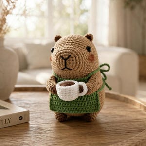 Peut inclure: Une figurine de capybara en crochet beige portant un tablier vert et tenant une petite tasse blanche. Le capybara a des yeux marron foncé, un nez et une bouche noirs brodés, et de petites marques de blush rose sur les joues en mailles visibles.