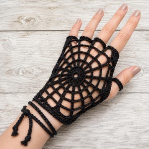 May include: A black crochet fingerless glove featuring a circular spider web pattern that covers the back of the hand. The design includes a matching ring loop for the middle finger and a long, thin cord that wraps around the wrist, ending in two small knotted tassels.