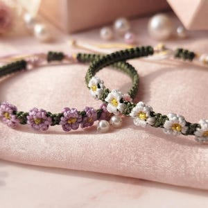 Könnte beinhalten: Zwei geflochtene Armbänder aus dunkelgrüner Kordel. Ein Armband zeigt eine Reihe kleiner, gehäkelter violetter Blumen mit gelben Mitten, während das andere eine Reihe weißer gehäkelter Blumen mit gelben Mitten und kleinen weißen Perlen aufweist.