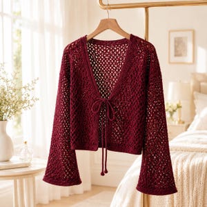 May include: A deep burgundy open-front cardigan featuring a loose crochet knit pattern, long flared sleeves, and a thin tie closure at the center of the neckline. The garment is constructed with a V-neckline and a cropped length, hanging on a hanger.