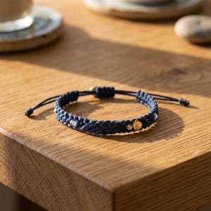 May include: A navy blue braided cord bracelet featuring small clear beads and a circular gold-toned charm engraved with the letter K. The bracelet has adjustable cord ends and a woven texture throughout the band, creating a simple circular design.