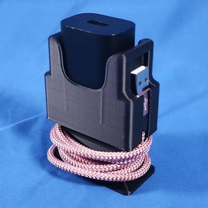 May include: A black plastic wall charger holder featuring a vertical slot for a power adapter and a side cutout for a USB port. A braided pink cable is coiled neatly around the base of the holder, which displays a small white USB icon symbol.