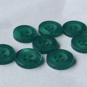 May include: A collection of seven round, translucent emerald green buttons. Each button features four central holes for attachment and the word GUCCI embossed in capital letters along the outer edge of the circular rim.