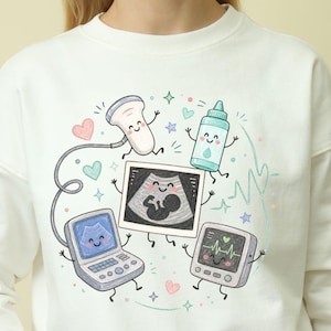 May include: A cream-colored crewneck sweatshirt featuring a colorful graphic illustration of anthropomorphic medical equipment, including an ultrasound probe, a gel bottle, a monitor, and a sonogram image, all with smiling faces.
