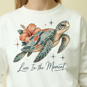 May include: A cream-colored crewneck sweatshirt featuring a graphic illustration of a sea turtle swimming near a large orange hibiscus flower. The design includes small star accents and the text "Live In the Moment" in a dark cursive script.