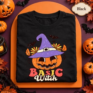 May include: A black t-shirt featuring a graphic of a smiling jack-o'-lantern wearing a purple witch hat with a white band and yellow flowers. The text "BASIC Witch" is printed below the pumpkin in a retro, multi-colored font with bats above.