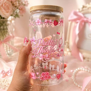 Puede incluir: Un vaso de cristal transparente con tapa de bambú que presenta estampados gráficos en rosa de lazos, flores, fresas, corazones y un pequeño bolso. El texto Pretty Things está impreso en el centro en una fuente rosa rodeado de elementos decorativos.