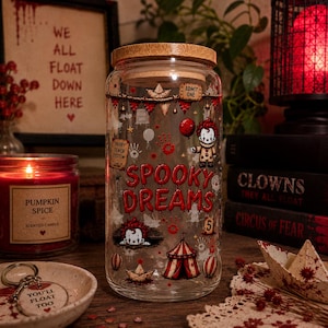 Puede incluir: Un vaso transparente con tapa de bambú que presenta un estampado gráfico colorido de un payaso, una carpa de circo, barcos de papel y el texto rojo "SPOOKY DREAMS". El diseño incluye pequeñas huellas rojas, un globo rojo y un ticket.