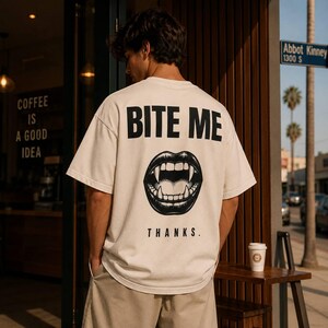 Könnte beinhalten: Ein cremefarbenes Kurzarm-T-Shirt mit einer schwarzen Grafik auf der Rückseite. Das Design zeigt den Text BITE ME oben, eine detaillierte Illustration eines offenen Mundes mit Vampirzähnen in der Mitte und das Wort THANKS. am unteren Rand.