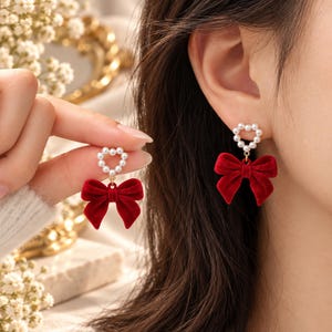 Può includere: Un paio di orecchini pendenti con un perno a forma di cuore realizzato con piccole perle bianche artificiali. Da ogni cuore pende un fiocco in velluto rosso intenso. Gli orecchini presentano un connettore in metallo dorato che unisce il cuore.