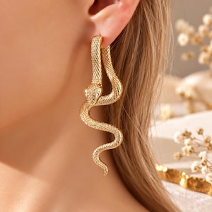 Puede incluir: Un pendiente dorado con forma de serpiente enroscada y una superficie texturizada similar a escamas. El diseño de la serpiente se curva hacia abajo desde el lóbulo de la oreja, con una cabeza detallada en la parte superior y un cuerpo fino.