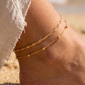 Op de afbeelding: Een set van twee goudkleurige enkelbandjes. Het bovenste enkelbandje heeft een gedraaid touwkettingontwerp, terwijl het onderste enkelbandje een fijne schakelketting is met kleine, gelijkmatig verdeelde goudkleurige kraaltjes.