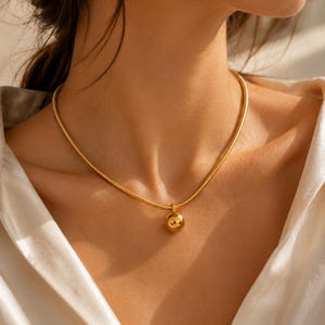 Puede incluir: Un collar de tono dorado que presenta un colgante esférico liso y pulido suspendido de una cadena de serpiente flexible. La superficie metálica refleja la luz, creando brillos intensos en el dije redondo y en los eslabones de la cadena.