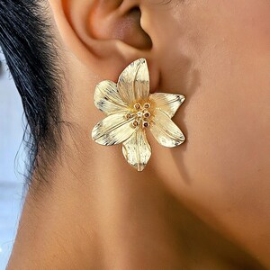 May include: A gold-toned metal earring shaped like a six-petaled flower. The petals feature a textured, linear finish radiating from the center, which is accented with small, dark brown circular stones arranged in a cluster at the core.