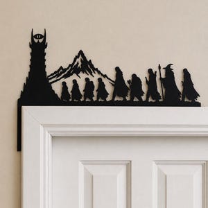 Op de afbeelding: Een zwarte metalen deurdecoratie met het silhouet van een hoge toren met een oog aan de top, een grillig gebergte en een groep van negen figuren die in een rij lopen, waaronder een figuur met een kap die een staf vasthoudt.