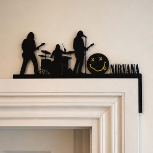 May include: A black metal door topper featuring a silhouette of a three-piece rock band with a drummer, a guitarist, and a vocalist. Next to the band is a yellow smiley face icon with X-shaped eyes and the word NIRVANA in black capital letters.
