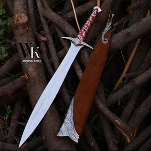 May include: A silver-toned metal dagger with a pointed blade and a red handle featuring white decorative patterns. The dagger is accompanied by a brown sheath with silver metal accents at the top and bottom. The text WARIVO KNIFE is visible.