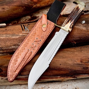 Peut inclure: Un couteau à lame fixe avec une lame en acier argenté, un manche en bois de cerf brun texturé et une garde en laiton. Il est accompagné d'un étui en cuir beige orné de motifs d'étoiles, de lacets et d'un bouton-pression en laiton. Le texte WK WARIVO KNIFE est visible.