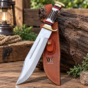 Peut inclure: Un couteau à lame fixe avec une lame en acier poli, une garde en laiton et un manche en bois de cerf texturé avec des accents rouges et noirs. Le couteau est accompagné d'un étui en cuir beige marqué du texte WK WAKVO KNIFE et d'un bouton-pression.
