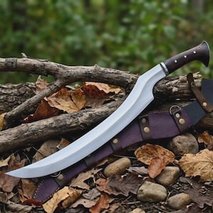 May include: A curved steel blade with a polished finish and a dark brown wooden handle featuring silver rivets. The knife is accompanied by a dark purple leather sheath secured with metal studs and rings, resting on a bed of fallen leaves.