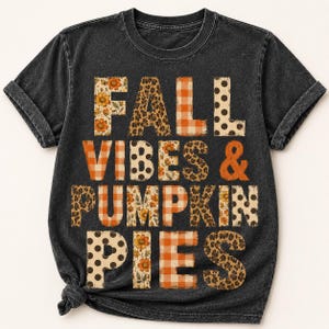 Pode incluir: Uma t-shirt de manga curta cinzento-carvão com decote redondo. A parte da frente apresenta o texto FALL VIBES & PUMPKIN PIES em letras grandes. Cada letra está preenchida com vários padrões, incluindo xadrez laranja, leopardo, bolinhas e flores.